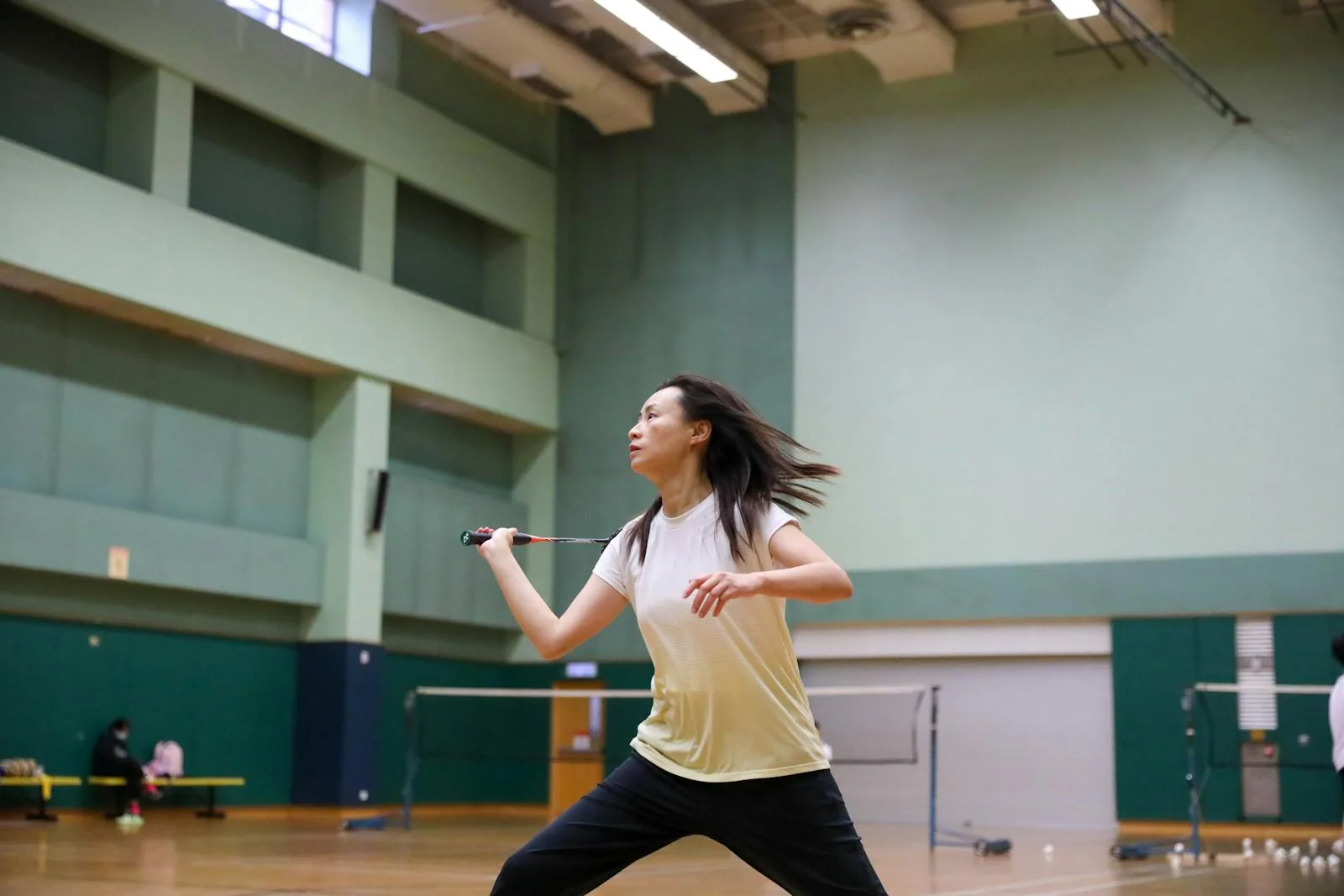 A player in a casual white shirt and black trousers in a ready stance mid-rally, in a green-walled municipal sports hall with a wooden floor.