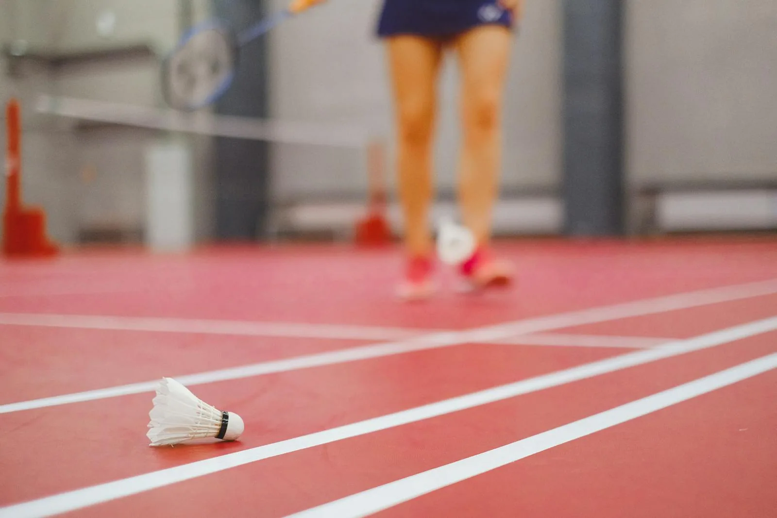 A feather shuttle resting on a court line with a blurred player in the background, after a rally.